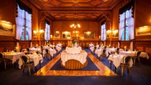 Elegant restaurantinteriør med trepaneler, lysekrone og hvite duker på runde bord, omgitt av malerier og gardiner.